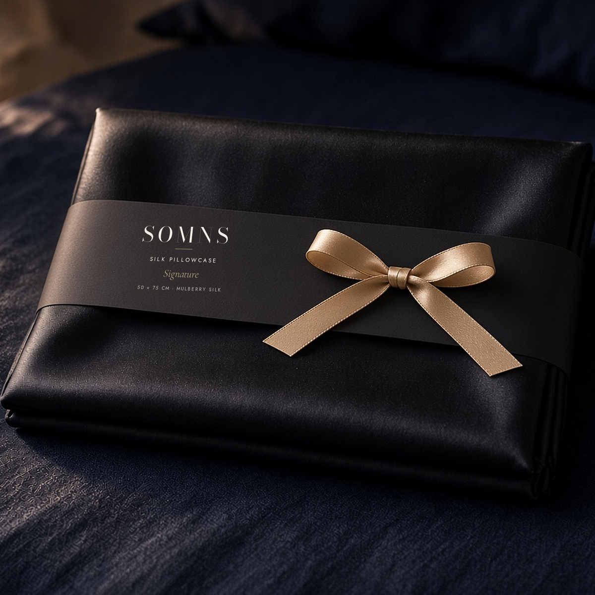 SOMNS black mulberry silk pillowcase, folded with a black SOMNS band and a champagne satin bow
