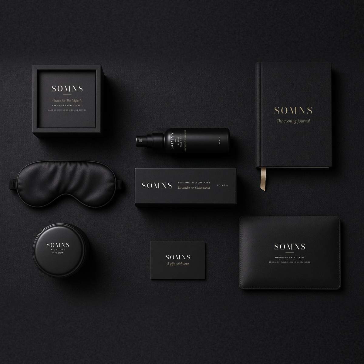 SOMNS The Night In gift set flat-lay on black: Bedtime Pillow Mist 50 ml and carton at the centre, with a glass candle in a black SOMNS box, silk eye mask, tea tin, bath flakes, evening journal and gift card